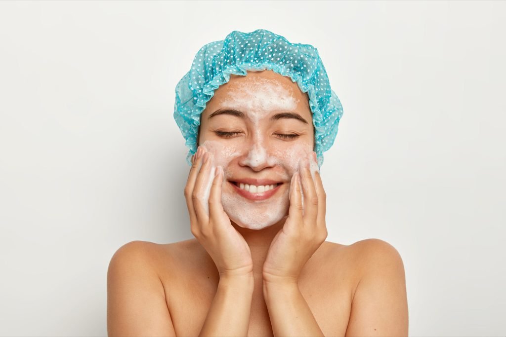 Portrait delighted young female model applies foaming facial cleanser touches cheeks has perfect fresh clean skin after sh...