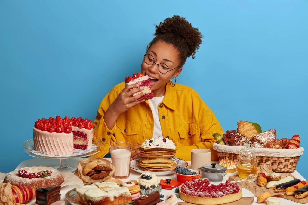 Hungry greedy afro american girl bites big delicious piece cake poses table with many yummy desserts has sweet breakfast h...