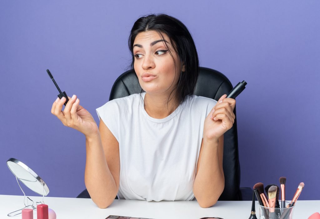 Impressed looking side beautiful woman sits table with makeup tools holding mascara