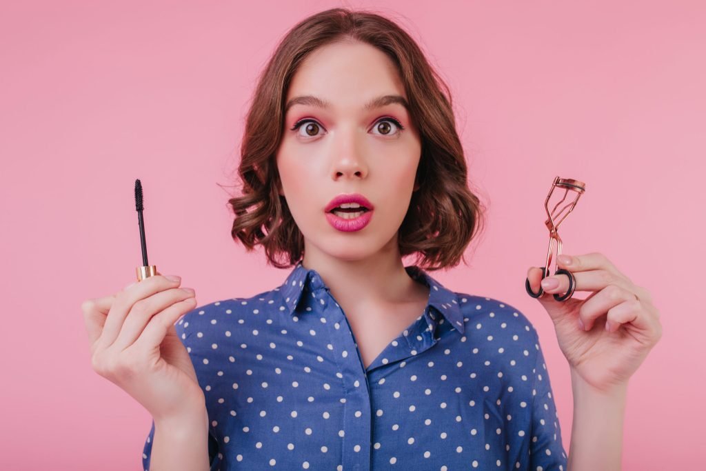 Magnificent lady with bright pink makeup posing with mascara indoor photo surprised cute girl curls her eyelashes