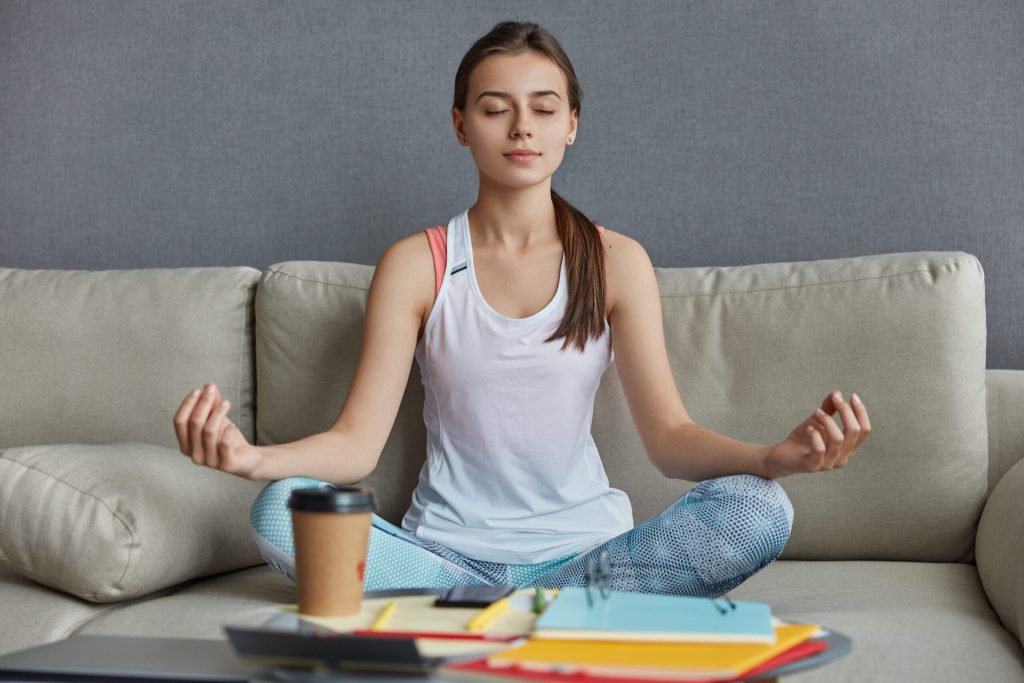 Relaxed concentrated teenager dressed casual clothes sits yoga pose meditates has break after hard work drinks takeaway co...
