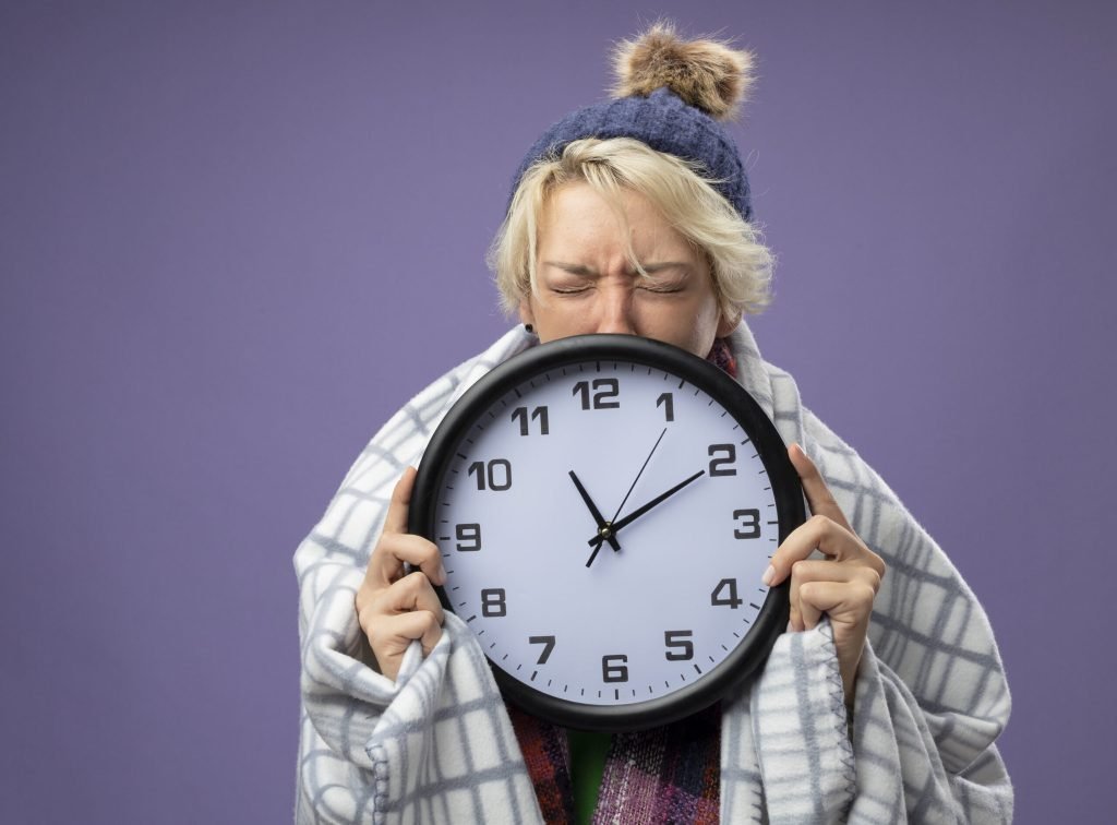 Sick unhealthy woman with short hair warm scarf hat feeling unwell wrapped blanket holding wall clock with closed eyes sca...