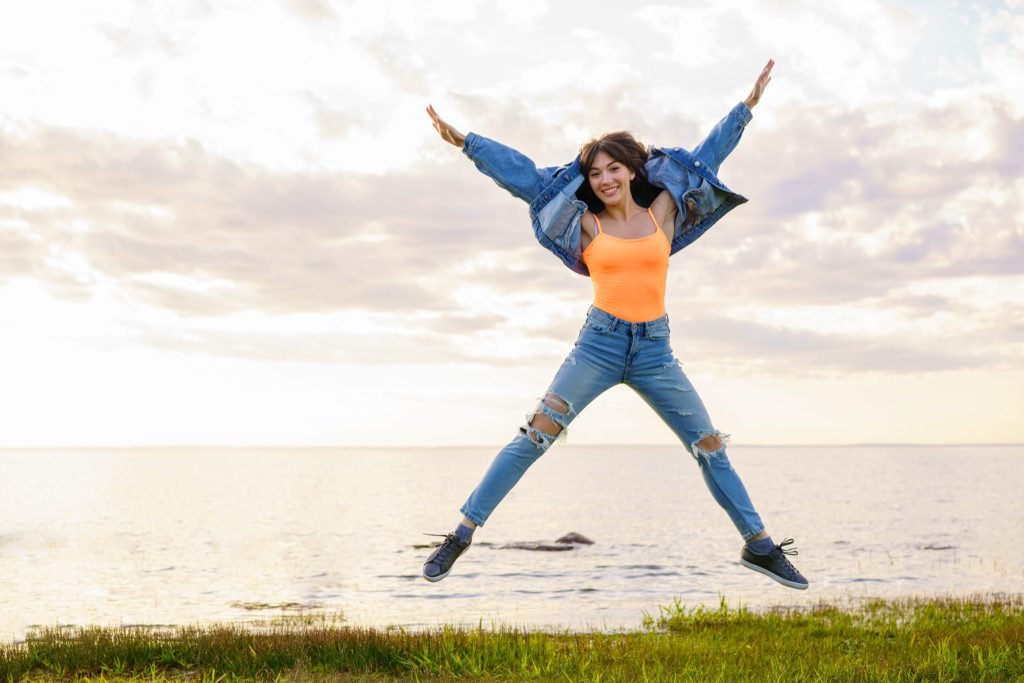 Young beautiful girl denim jacket jeans yellow t shirt jumps background sea summer day posing sunset