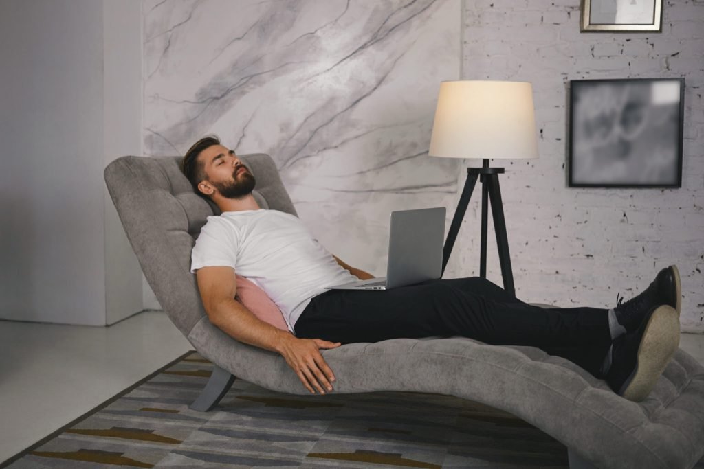 Candid shot unshaven young male shoes lying comfortably gray couch with portable computer his lap having nap meditating ke...