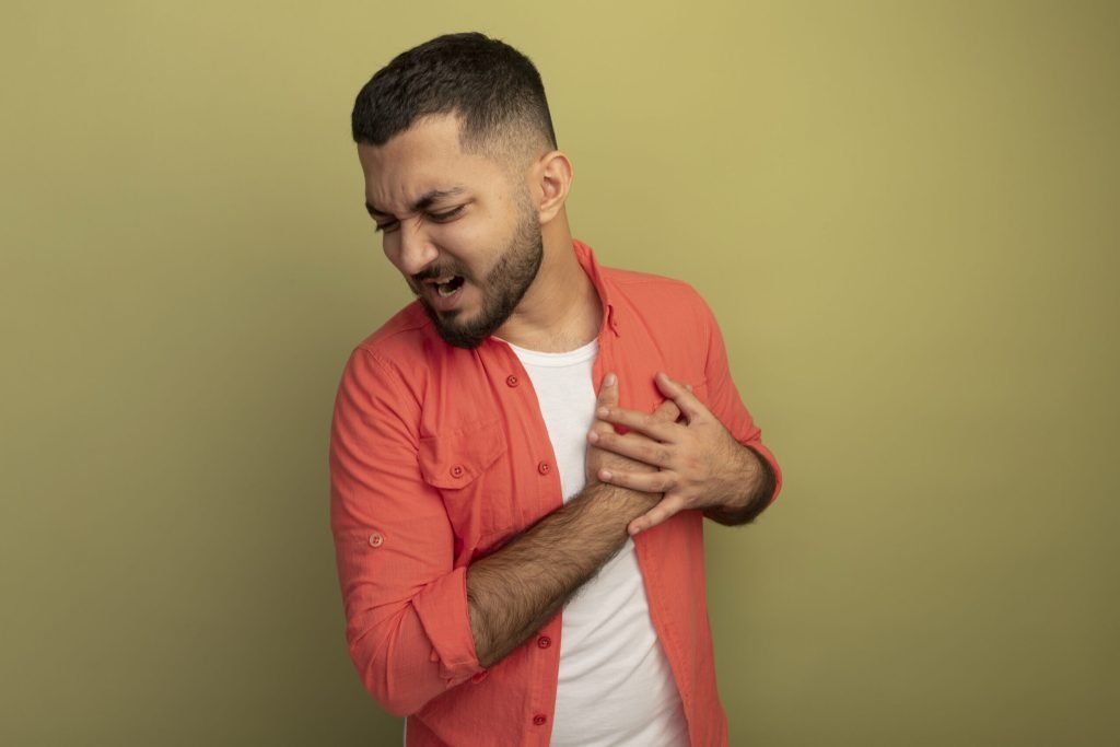 Young bearded man orange shirt holding hands his chest feeling pain standing light wall
