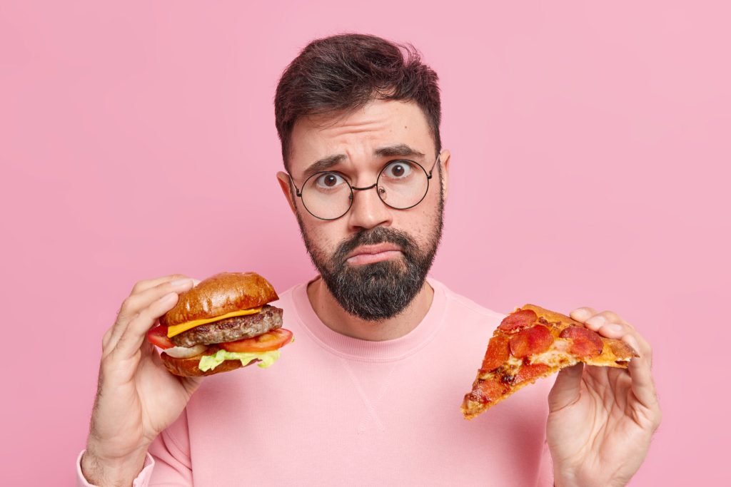 Photo displeased bearded man cannot refuse eating fast food holds delicious hamburger slice tasty pizza looks unhappily ha...