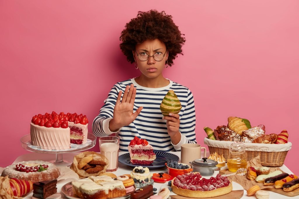 Serious displeased woman with afro hairstyle shows refusal gesture holds croissant denies eating dessert wears spectacles ...