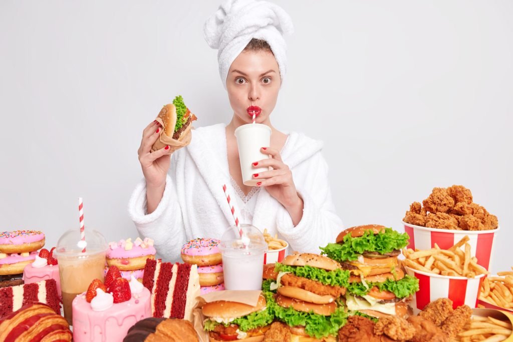 Unhealthy nuttrition concept housewife with red manicure lips domestic bathrobe towel head drinks soda eats junk food
