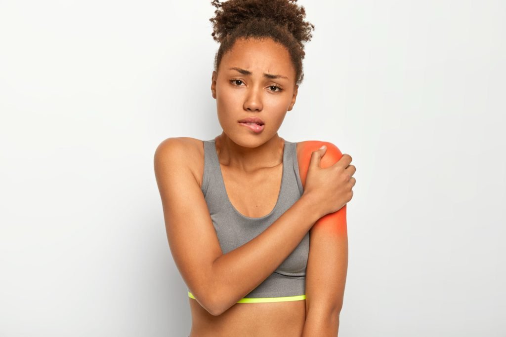 Displeased curly haired woman bites lips touches painful shoulder suffers from injury wears grey top isolated white backgr...