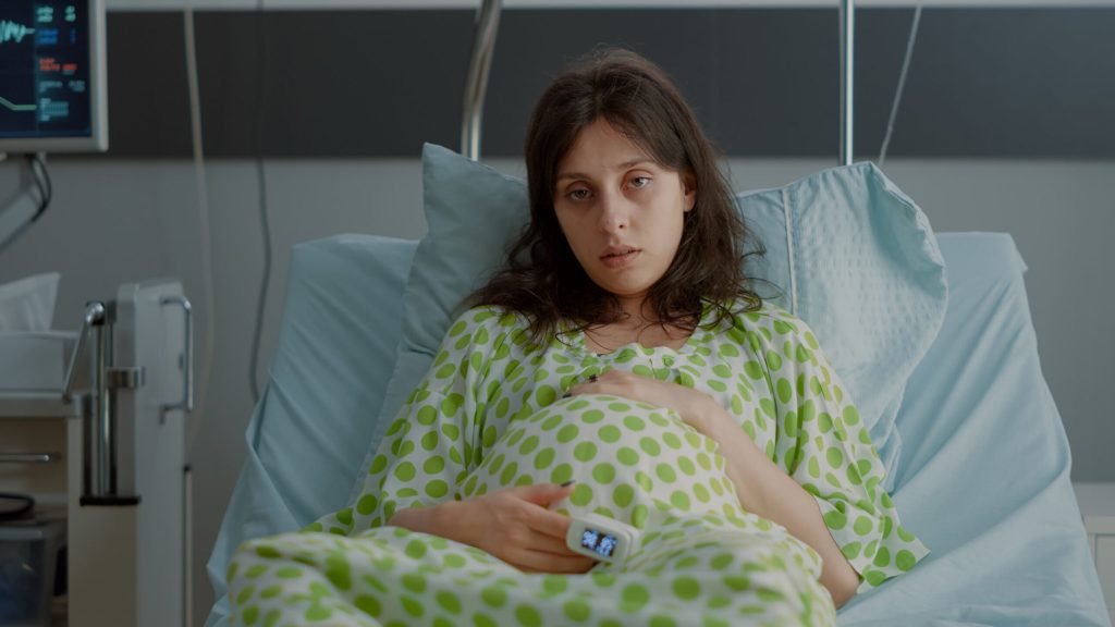 Tired caucasian woman with pregnancy belly sitting bed maternity room young pregnant person with oximeter feeling tension ...