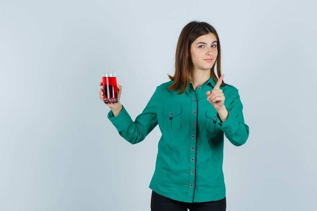 Young girl holding glass red liquid showing warning gesture green blouse black pants looking cute front view