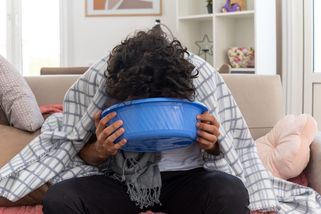 Sick young caucasian man optical glasses wrapped plaid with scarf around his neck holding vomiting into bucket sitting cou...