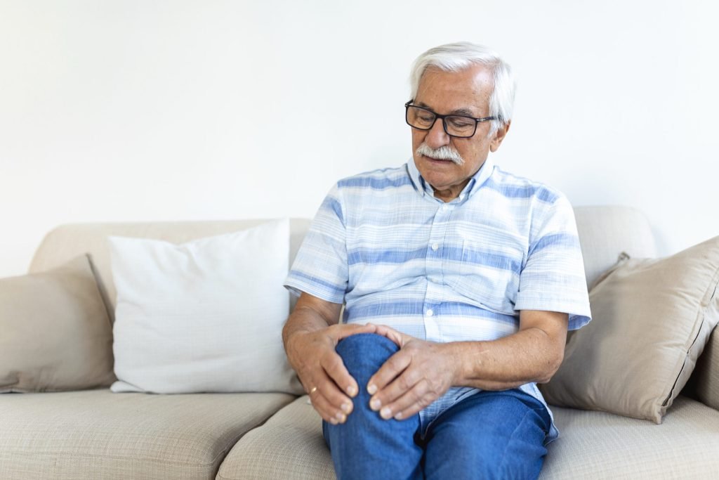 Elderly man sitting sofa home touching his painful knee people health care problem concept unhappy senior man suffering fr...