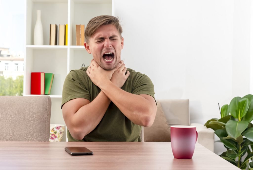 Annoyed young blonde handsome man sits table with phone cup choking himself with two hands inside living room