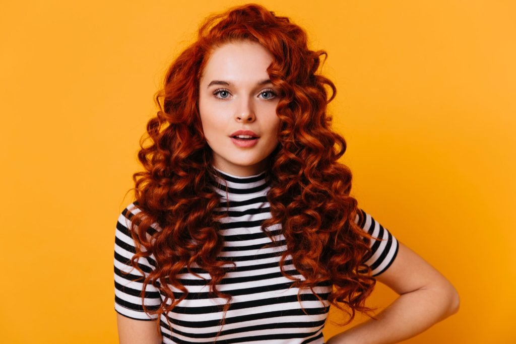 Close up portrait blue eyed girl with long eyelashes voluminous red curly hair against orange space