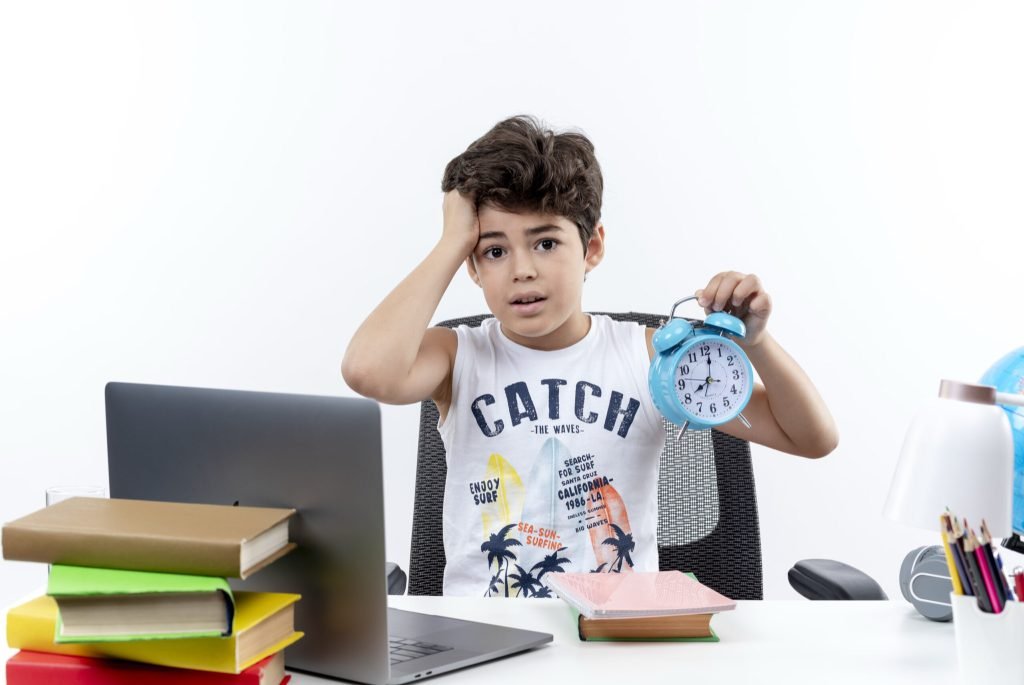 Concerned little schoolboy sitting desk with school tools holding alarm clock grabbed head isolated white background