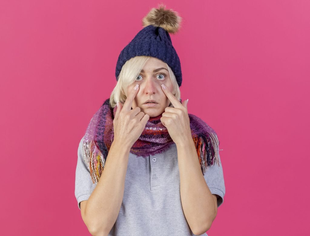 Surprised young blonde ill slavic woman wearing winter hat scarf pulls down eyelids isolated pink wall with copy space