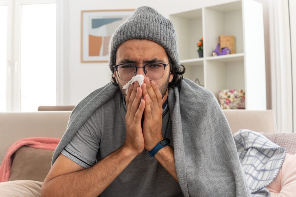 Unpleased young man optical glasses wrapped plaid wearing winter hat wipes his nose with tissue looking front sitting couc...