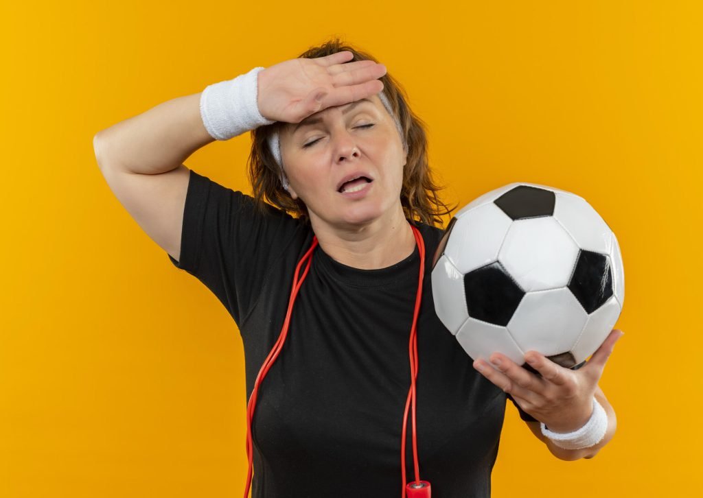 Middle aged sporty woman black t shirt with headband holding soccer ball looking tired overworked standing orange wall