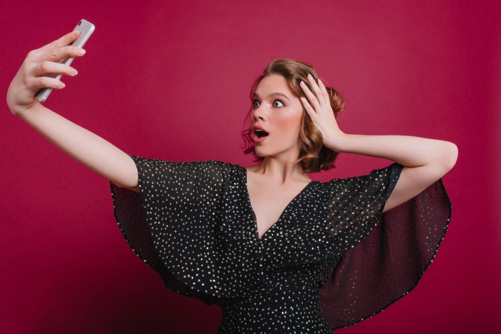 Amazed woman plays with short shiny hair while making selfie shocked caucasian girl vintage casual dress taking photo hers...