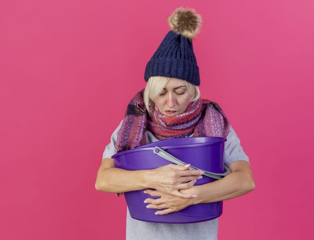 Unpleased young blonde ill slavic woman wearing winter hat scarf holds looks plastic bucket having nausea isolated pink wa...
