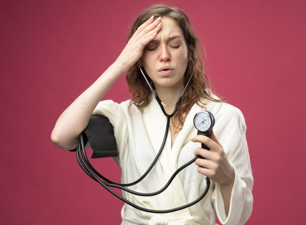 Weak young ill girl with closed eyes wearing white robe measuring her own pressure with sphygmomanometer putting hand fore...